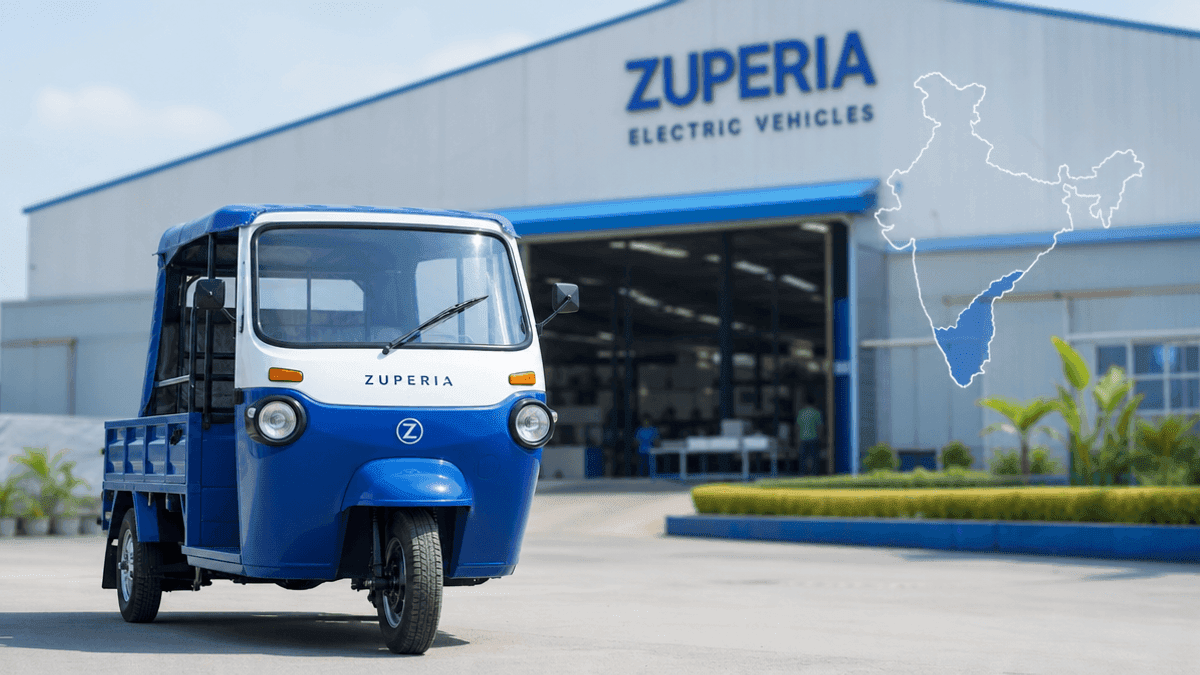 A Zuperia electric three-wheeler vehicle positioned in front of a factory building with a blurred industrial background.