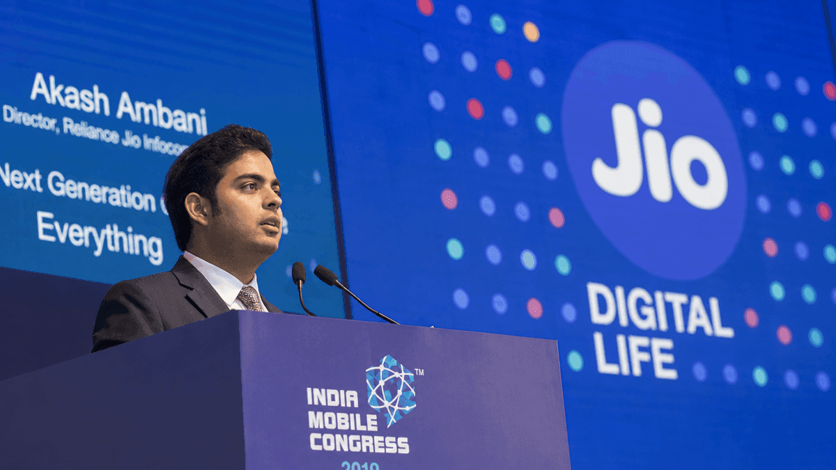 Akash Ambani speaking at a podium at the India Mobile Congress with a large screen displaying Jio branding behind him.